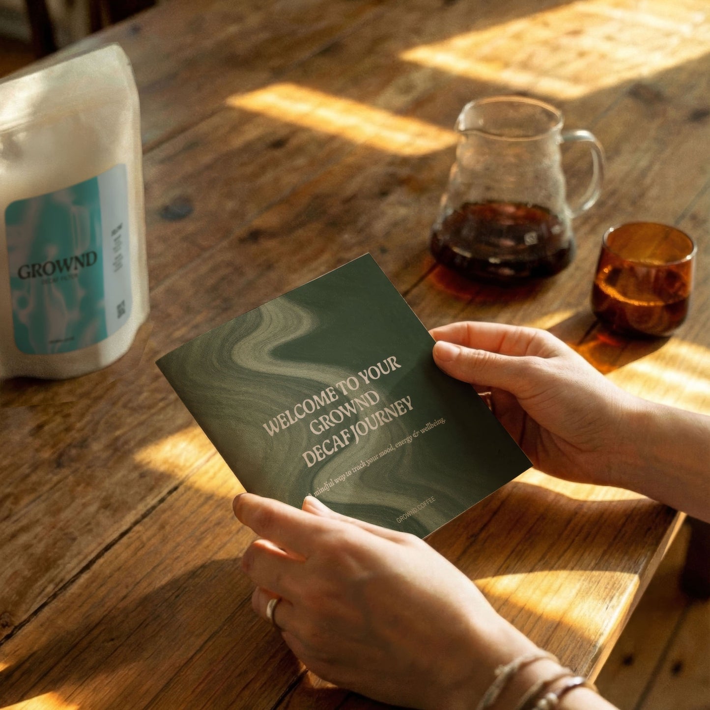 Person hält eine GROWND Mood Tracker auf einem Holztisch neben einer Glaskanne mit Kaffee und einer Kaffeeverpackung im warmen Sonnenlicht.