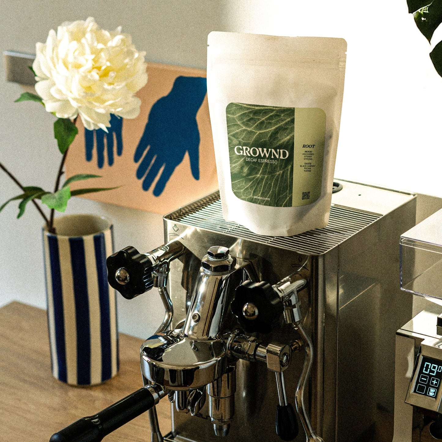 Espresso machine with a bag of Grownd coffee on top, next to a vase with a flower and a piece of art.